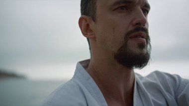 Kimono bulutlu bir sabahta karate eğitimi alan ciddi bir orta yaşlı adam. Güçlü sporcu elleri sahilde dövüş egzersizleri yapıyor. Odaklanmış sakallı sporcu dışarıda dövüş sanatları yapıyor..