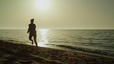 Yaz günbatımında arka manzara koşucusu kadın. Spor kıyafetleriyle deniz kıyısında koşan tanınmamış atletik kız. Aktif eğitimli sporcu kadınla güzel bir akşam manzarası. Doğa konsepti üzerinde sağlıklı spor.