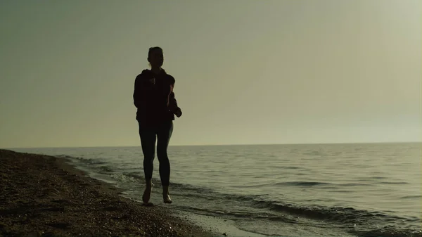 Silhouette of athletic woman running sandy beach at summer sunset. Sporty girl jogging on seacoast washed ocean waves. Confident sportswoman exercising wearing sportswear on nature. Healthy lifestyle