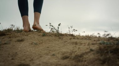 Tanınmayan sporcu kız tepenin kenarına gidiyor ve güzel deniz manzarasının tadını çıkarıyor. Yeşil çimen tepesinde yürüyen çıplak ayaklı bir kadın bulutlu bir günde. Spor kadını ayakları yüksek tepeciklere tırmanıyor.. 