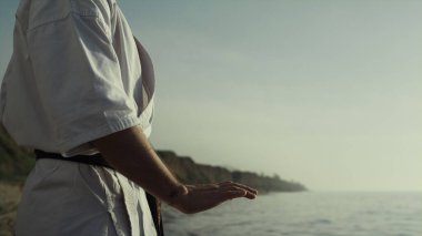 Bilinmeyen karate ustası deniz kıyısında dövüş egzersizleri yapıyor. Atletik adam gün batımında meditasyon tekniği eğitimi alıyor. Güçlü sporcu elleri doğada dövüş sanatları uyguluyor.. 