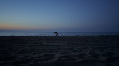 Siluet sportif kız gecenin geç saatlerinde plajda yoga yapıyor. Alacakaranlıkta tanınmayan sporcu kadın eğitimi. Atletik kadın deniz kıyısında esneme hareketi yapıyor. Spor sağlık konsepti. 