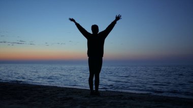 Bilinmeyen yogi kadın deniz kıyısında güzel bir akşamda rahatlatıcı egzersizler yapıyor. Siluet, koyu mavi günbatımı gökyüzünün önünde egzersiz yapan bir kız. Sıska sporcu kadın, el kaldırıyor, dışarıda antrenman yapıyor.. 