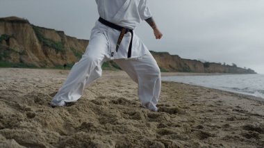 Erkek bacakları kumlu sahilde karate egzersizi yapıyor. Dışarıda kasvetli bir günde dövüş sanatları yapan tanınmamış bir dövüşçü. Güçlü sporcu deniz kenarındaki bulutlu sabahlarda dövüş eğitimi alıyor.