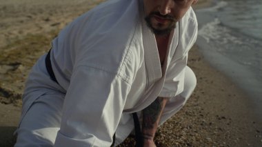 Kumsalda kimono giyerek esneme hareketleri yapan sportif bir adam. Sakallı sporcu antrenman dengesi açık havada. Deniz kıyısında yaz günü karate yapan güçlü bir atlet. Aktif sağlıklı yaşam tarzı.