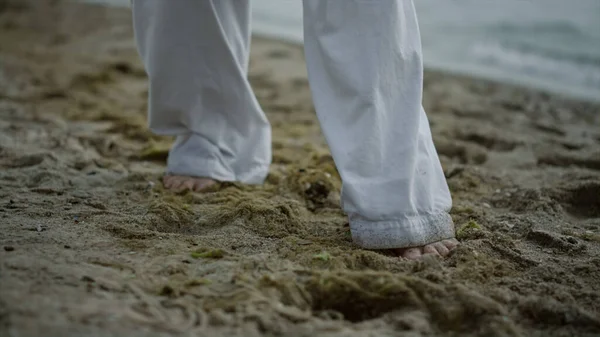 Unknown barefoot man stepping on sandy beach close up. Young athlete exercising on wet seacoast outdoors wearing white sportswear. View legs unrecognizable sportsman training karate summer morning.
