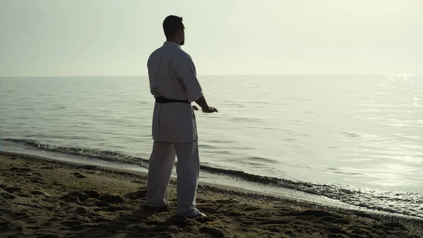 Calm barefoot man training karate watching picturesque sunset on beach. Focused fighter practicing martial arts in sunlight standing sand seacoast. Brutal sportsman exercising on nature wearing kimono