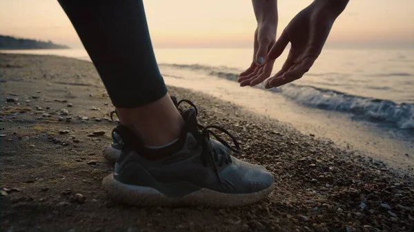 Unknown sportswoman reaching hands to ground standing on wet sand outdoors close up. Sporty woman bending body forward stretching at sunset. Unrecognizable slim girl training near ocean waves.