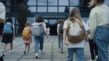 Diverse children going school with backpacks back view. Cheerful smiling teachers meet welcoming teen students at open door. Excited elementary age pupils entering modern building. Study concept.