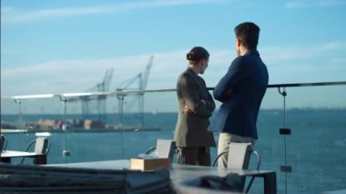 Couple looking sea terrace in morning sunlight. Business partners enjoy view resting on rooftop cafe at shipping port cranes. Commercial managers relax talking on break discussing financial project.