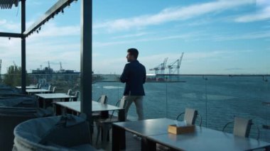 Successful executive going terrace in luxury cafe. Man enjoying calm sea view resting alone. Millennial manager freelancer walking glass balcony relaxing in morning. Confident business owner concept.