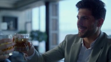 Handsome gentleman clinking glass in restaurant closeup. Businessman drinking cocktail at dinner in fancy panoramic window cafe. Leader person portrait tasting bourbon beverage with unknown colleague