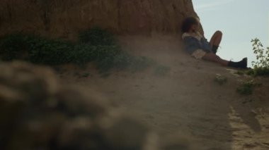 Kaygısız stil kızı gölgede oturup güneşli sonbaharda plaj tepesine yaslanıyor. Çekici kıvırcık kadın kumsalda rahatlıyor. Güzel doğada poz veren kendinden emin genç bir model. Boş zaman konsepti.