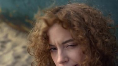 Güzel bir kız kameraya benziyor. Kıvırcık saçlarını yakından çekiyor. Çekici genç bir kadın eski teknenin yanında oturmuş kıvırcık saçlarını sallıyor. Deniz kıyısında yalnız poz veren neşeli kadın kasvetli bahar sabahı.