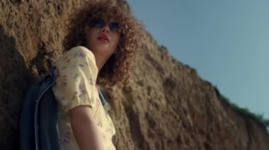 Cazibeli model deniz kıyısında güneş ışığını yakıyor. Güneş gözlüklü, şık bir elbise giymiş kıvırcık, çekici bir kadın. Kendine güveni tam genç bir kız. Kum tepesine yaslanmış rahatlıyor..