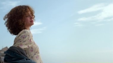 Sahilde dikilen çekici kız, gün ışığına karşı yüzünü yukarı kaldırıyor. Güzel, mavi gökyüzünün önünde kıvırcık saçlarını sallayan sevimli, mutlu, rahat bir kadın. Hayalperest kadın, güneşli deniz manzarasının tadını çıkar..