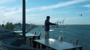 Young businessman resting outdoors enjoying sea view on glass cafe terrace. Handsome business owner lean thinking career work problems at port cranes. Successful millennial guy relaxing in restaurant.