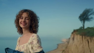 Portrait of happy young lady smiling on beautiful beach. Attractive curly girl looking camera touching face with hand. Cheerful relaxed woman standing empty seashore wearing jeans jacket. Rest concept
