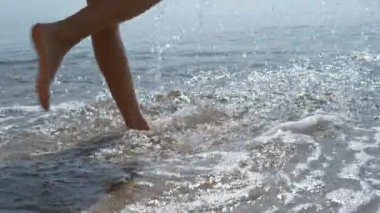 Young girl feet jumping on sea waves close up. Unknown carefree barefoot woman playing splashing ocean water on summer sunlight. Slim tanned lady legs stepping on seashore sunny day. Vacation concept.
