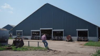 Çiftlik işçisi çiftlik hayvanlarını besledikten sonra çitin üzerinde büyük bir ahırda oturuyor. Yorgun tarımsal işletme sahibi ineklerle çiftçilik tesisinde çok çalışıyor. Modern tarım kompleksi inşa kavramı.