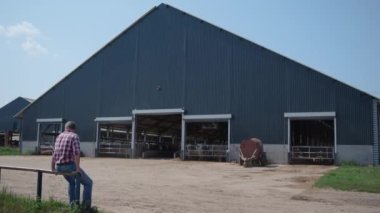 Çiftçi, çiftlik çitlerinde oturmuş modern çiftlik hayvanı üretiminde sıkı çalıştıktan sonra dinleniyor. Profesyonel çiftlik işçisi büyük ahır binasının önünde dinleniyor. Ekolojik organik tarım kavramı.