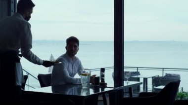 Relaxed man resting hotel cafe terrace at lunch time break. Elegant masked waiter serving meal to successful businessman customer. Joyful entrepreneur silhouette enjoying sea view in luxury restaurant