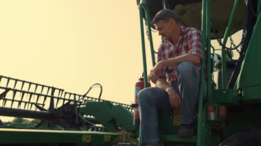 Farmer resting combine machine at countryside. Driver tractor checking equipment at grain farmland. Tired worker farmer planning future harvesting after hard working day. Agritech specialist concept.