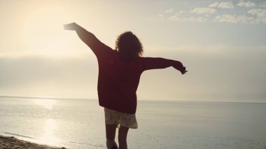 Gündoğumunda deniz kıyısında yürüyen sıradan bir kız. Pozitif kadın el kaldırıyor. Başarıyı kutlayan kadın. Genç bayan özgürlüğün tadını çıkarıyor. Güneş ışıkları okyanus yüzeyine düşüyor. Zafer konsepti