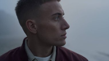 Kapalı çekim odaklı adam yüzü dışarıdaki kameraya bakıyor. Sisli bir sabahta sahilde poz veren yakışıklı adamın portresi. Erkek model baştan başa dönüyor. Ciddi bir adam başka tarafa bakıyor. Deniz kıyısında duran kişi