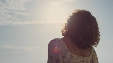 Bilinmeyen kıvırcık kadın, yüzünü güneş ışığına çeviriyor. Arka manzara genç, rahat kız dışarıda tek başına parlak sabah güneşinin tadını çıkarıyor. Doğada rahatlayan sevimli elbiseler giyen tanınmayan bir kadın..