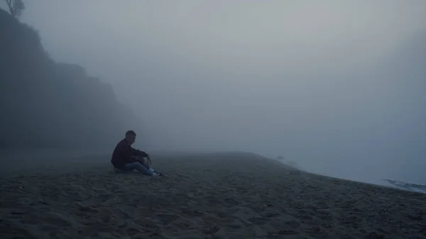 Wide shot depressed guy sitting on beach in foggy morning. Worried man touching sand grains. Male hipster feeling loneliness. Man looking ocean landscape. Misty atmosphere. Autumn scenery at sea coast