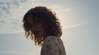 Muhteşem kıvırcık kız, parlak güneş ışığının önünde kameraya bakıyor. Doğada poz veren genç ve çekici bir manken. Güzel elbiseler giyiyor. Deniz kıyısında tek başına duran gülümseyen güzel kadına yakın dur. Doğal güzellik..