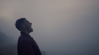 Yakın plan, gün doğumunda deniz sahilinde yürüyen üzgün adam. Dışarıda çığlık atan bitkin bir adam. Sisli hava. Erkek model stresten kurtuluyor. Okyanus kıyısında sisin içinde duran üzgün adam. Ruh sağlığı konsepti 
