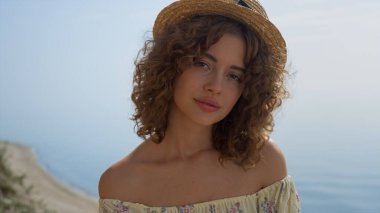 Kıvırcık, nazik bir kadın kameraya bakıyor. Önünde durgun bir gülümseme var. Güneşli deniz kıyısında poz veren hasır şapkalı çekici kızın portresi. Genç bayan doğada rahatlayın..