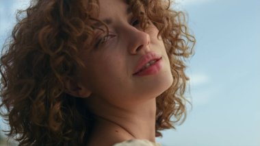 Kumlu kumsalda güneşli bir günde çiçekli elbiseler giyen hoş bir bayan. Kapatın. Deniz kıyısında tek başına duran çekici kıvırcık kadın deniz manzarasının tadını çıkarıyor. Zarif, baştan çıkarıcı bir kız, sakin bir gülümsemeyle poz veriyor..