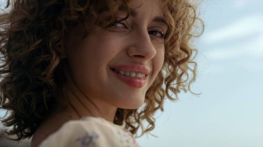 Mavi, güneşli gökyüzünde gülümseyen güzel bir kadın yüzü. Yaklaş. Deniz sahilinde güzel kıvırcık saçlı neşeli çekici bir kızın portresi. Güzel elbiseli kadın, baştan çıkarıcı bir şekilde kameraya bakıyor..