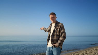 Kareli gömlekle güneşli deniz kıyısında poz veren gülümseyen adam. Çekici genç adam güzel deniz manzarasına el kaldırarak video blogu hazırlıyor. Havalı mutlu blogcu kumlu okyanus sahilinde dinleniyor..