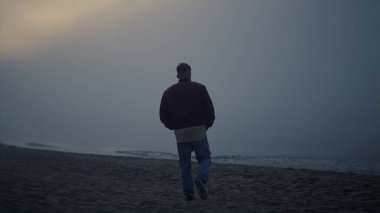 Sabaha karşı kumlu sahilde yürüyen genç adam. Deniz ufkuna bakan yalnız bir adam. Erkek model okyanus kıyısında yürüyor. Barışçıl adam güneşin doğuşunu seyrediyor. Depresyon ve üzüntü konsepti