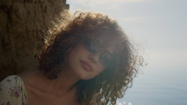 Güneş gözlükleriyle kumsalda güneşli bir günde saçlarını sallayan gülümseyen kıvırcık kızın portresi. Mavi okyanusun önünde poz veren mutlu rahat model parlak güneş ışığını yaktı. Çekici, stil sahibi bir kadın..