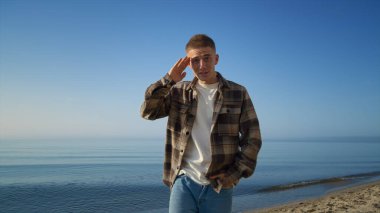 Güzel deniz kıyısında güneşli bir sabahta kameraya yaklaşan mutlu stil sahibi adam. Genç yakışıklı, doğa üzerine konferans salonuyla iletişim kuruyor. Okyanus sahilinde seyahat blogu yazan pozitif adam..