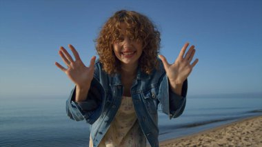 Güzel kumsalda kameraya bakan sevimli, çekici bir kadın. Yakından. Deniz kıyısında el sallayarak video bloğu çeken gülümseyen genç kızın portresi. Şık kot ceket giyen neşeli, güzel bir manken..