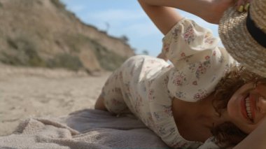 Sahilde oynayan, hasır şapkayla kamerayı örten pozitif sakin kız. Neşeli bir kadının portresi güneş ışığında kumsalda yatıyor. Şirin elbiseli çekici genç bayan yaz doğasında rahatla..
