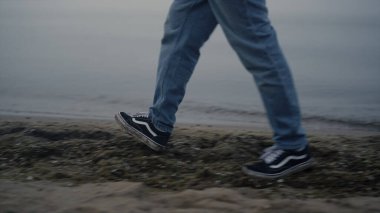 Deniz kıyısında yürüyen, sıradan spor ayakkabılı, tanınmayan bir adam. Erkek ayakları kumlu sahilde yürüyüşün tadını çıkarıyor. Kot pantolon giyen bir erkek model. İnsan bacakları sisli bir sabahta kıyı şeridi boyunca doğayı keşfediyor.