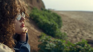 Güzel güneş gözlüklü çekici kız yüzüne yaklaş. Kum kıyısında tek başına oturan rüya gibi rahat kadın portresi. Kıvırcık genç bayan güzel sonbahar deniz manzarasının tadını çıkarıyor..