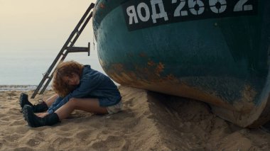 Gevşemiş genç bayan, boş deniz kıyısında vücut bacağa büküyor. Çekici kıvırcık kadın sahilde eski terk edilmiş teknenin yanında dinleniyor. Açık havada poz veren rüya gibi ciddi bir kız kot ceket giyiyor. Hafta sonu konsepti