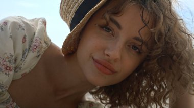 Güzel, gülümseyen bir kadın güneş ışığından saklanan hasır bir şapkayla yüzünü kapatıyor. Sahilde güneşli bir hafta sonu geçiren çekici mutlu bir kadının portresi. Neşeli kıvırcık kız neşeyle kameraya bakıyor