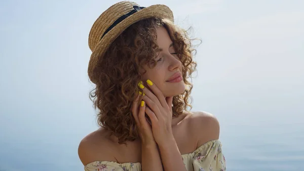 Seductive woman closed eyes enjoy sunlight on beautiful seacoast close up. Portrait of attractive curly girl wearing straw hat gentle touching face. Relaxed young lady posing on sunny summer beach.