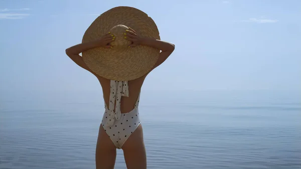 Relaxed stylish girl standing beach raising hands to wide brimmed hat close up. Back view young slim lady looking amazing seascape at summer sunlight. Carefree woman in swimsuit enjoy sunny seacoast.