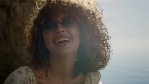 Laughing girl shaking head sitting beach in front blue ocean close up. Curly curls bouncing merrily lighted soft summer sunlight. Portrait of attractive smiling woman sitting seashore in sunglasses.