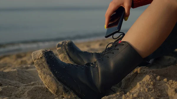 Stylish girl sitting on beach at vacation. Unrecognizable woman hand holding mobile smartphone. Woman hipster legs wearing casual boots. Leathers shoes in sand. Ocean waves splashing focus on shore.